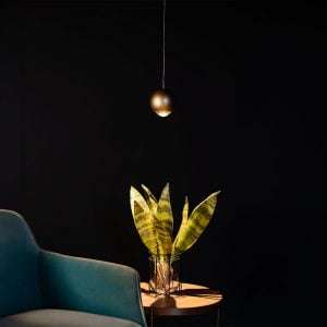 Um ambiente escuro, fundo preto, uma parte de uma poltrona verde azulada aparecendo ao canto esquerdo, uma mesinha de madeira ao centro com um vazo de vidro com folhas longas dentro, acima há o pendente Muna iluminando a mesa e a planta.