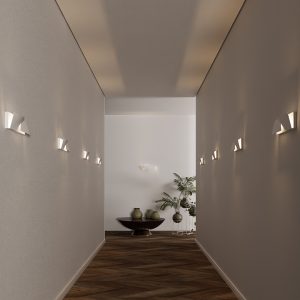 A imagem mostra um corredor moderno, com piso de madeira em padrão espinha de peixe e paredes lisas em tom claro. Nas laterais, há várias arandelas de parede instaladas em linha reta, com design geométrico e luz indireta, projetando feixes para cima e para baixo. A iluminação cria um efeito suave e elegante no ambiente, destacando a arquitetura do espaço. Ao fundo, vê-se uma mesa baixa com vasos decorativos e uma planta, completando a composição com um toque natural. O conjunto transmite sofisticação e aconchego, com um estilo minimalista e bem equilibrado.