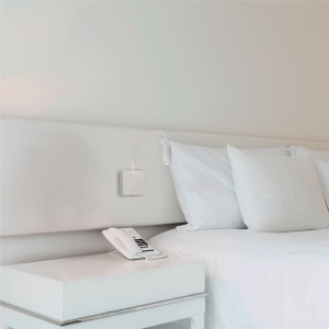 White hotel bed with two pillows, wall-mounted curved reading lamp, and a phone on a white bedside table.