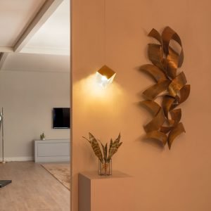 Um ambiente quente devido a luz amarelada do pendente BITIS que está no centro bem próximo da parede branca de fundo, permitindo ver o efeito que o seu vidro texturizado da a luz que reflete na parede. Na parede, ao lado direito também há uma escultura feita de laminas de metal curvas na cor cobre. Abaixo do pendente há uma mesinha cubica cinza claro, com um vazo de vidro com folhas longas dentro. Ao lado esquerdo a parede acaba logo após a mesinha, permitindo ver ao fundo um ambiente mais claro onde há um piso de madeira, um tapete cor caramelo, um móvel cinza com uma porta e um pequeno vazo de planta em cima e uma TV presa a parede acima do móvel.