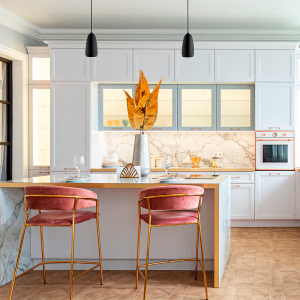 Modern kitchen with marble island, pink velvet bar stools, and white cabinets; orange leaf centerpiece.