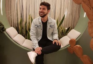 Young man with a denim jacket sits crosswise on a curved green bench with white cushions, smiling in a plant-filled indoor setting.