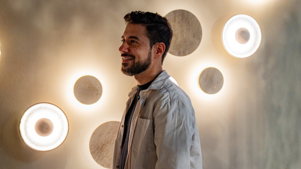 Smiling man with dark hair and a trimmed beard stands next to a wall of round, glowing discs.