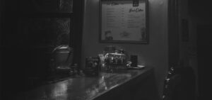 Dimly lit grayscale cafe counter with a framed menu board and a glass beverage dispenser on the counter.